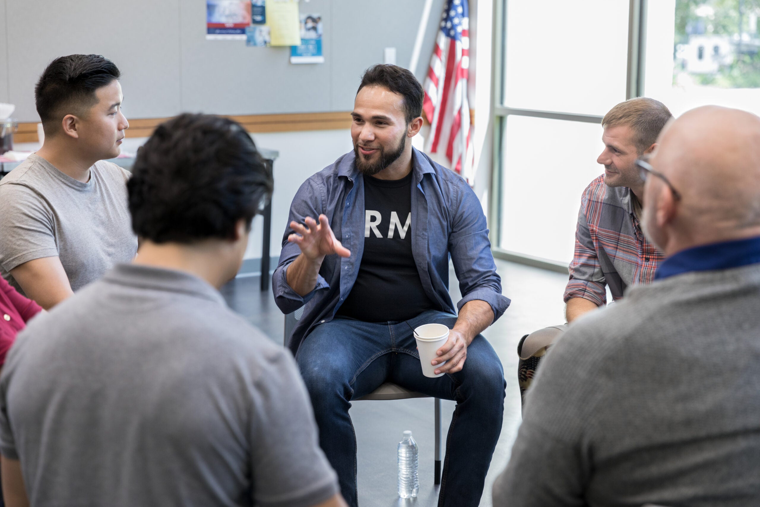 People seated in a circle listen as a man gestures while holding a paper cup.