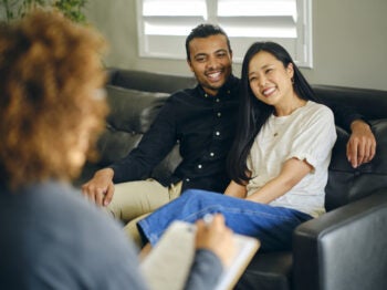 Couple seated on a sofa speaking with a counselor holding a clipboard.