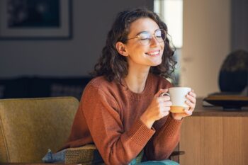 Smiling woman in glasses holding a white mug while seated indoors.