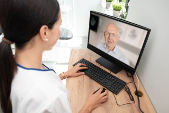 Person in a white coat uses a computer to speak with an older man on a video call.