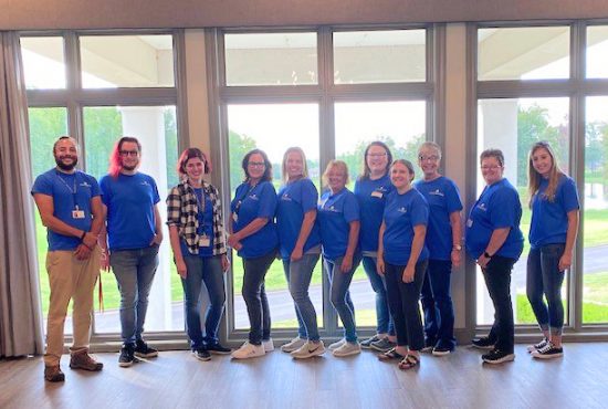 Ten adults stand in a row indoors in front of large windows, most wearing blue shirts and lanyards.